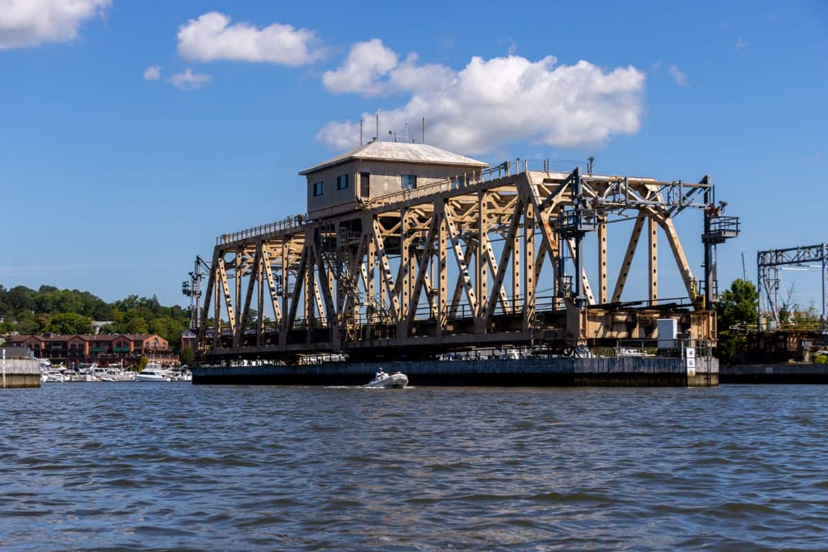 A steel truss drawbridge with a central control house spans a river under a blue sky; a small boat passes underneath.