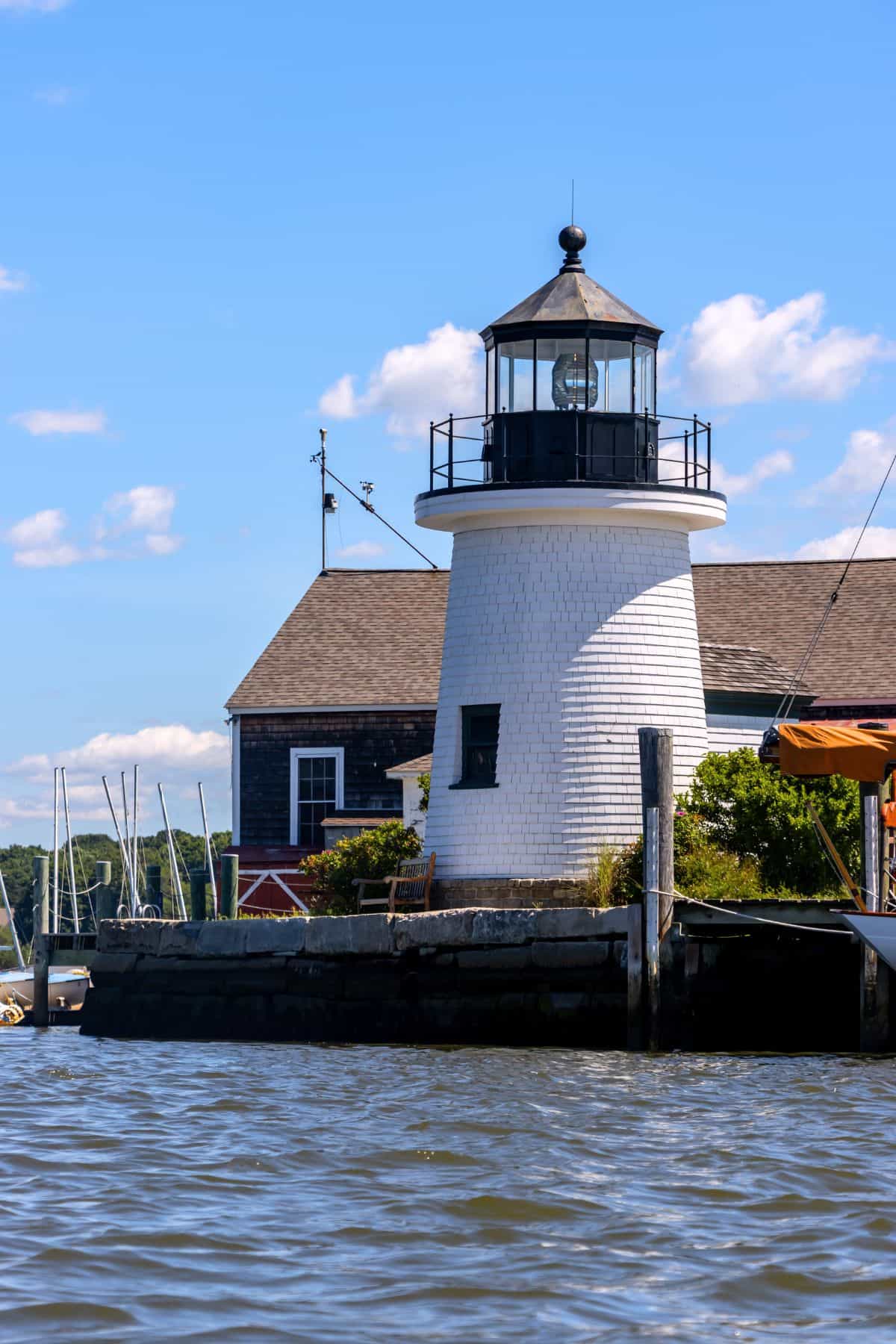 White lighthouse with a black lantern sits on a dock next to a wooden building under a blue sky with scattered clouds, viewed from the water.