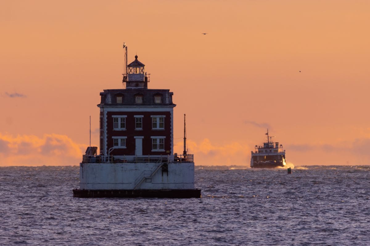 Square-shaped lighthouse on the water at sunset with a ferry approaching in the background under an orange sky.