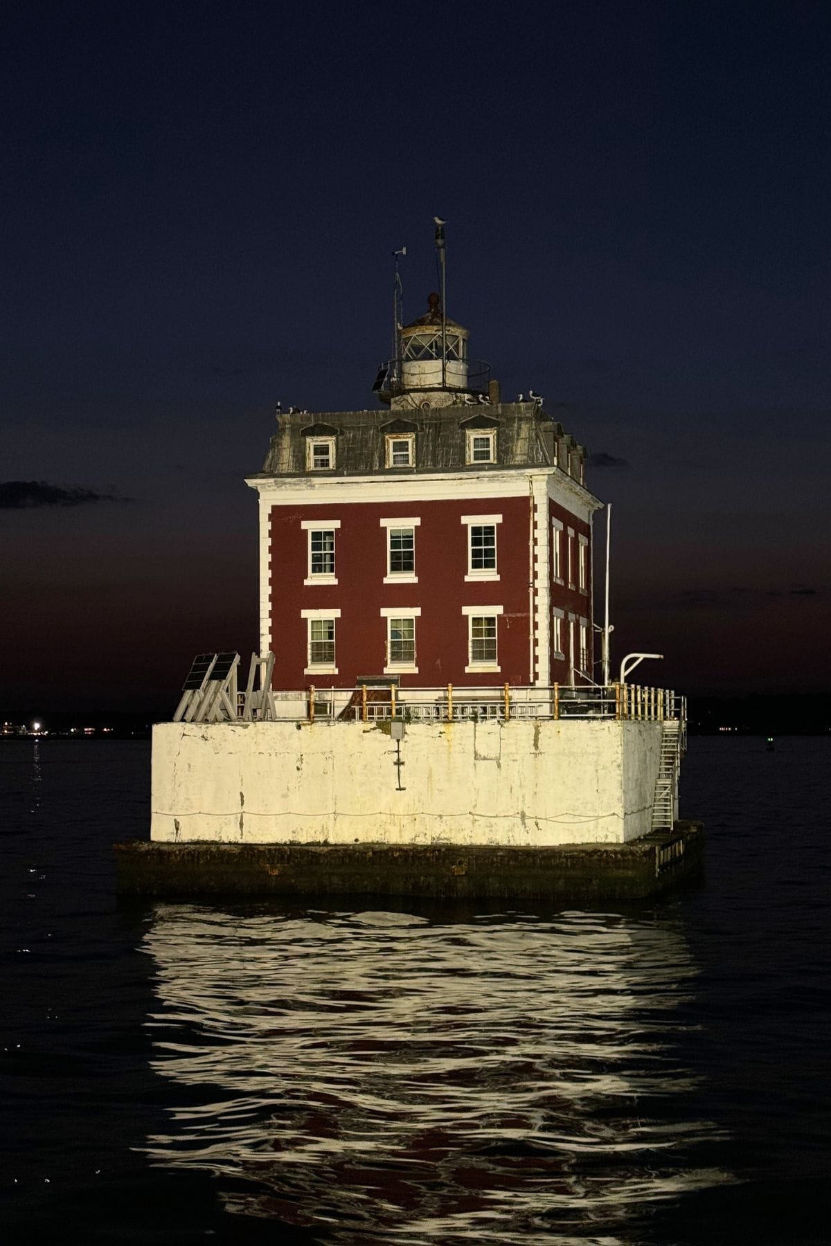 A square, red-brick lighthouse with white trim sits on a concrete platform surrounded by water at dusk, with its reflection visible on the surface.