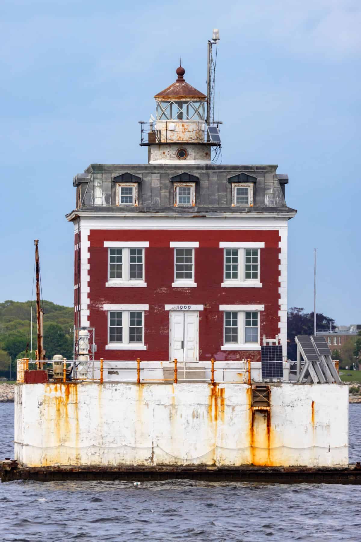 A square, red-brick lighthouse with a white base and rust stains stands in the water, featuring a lantern room on top and several windows on its facade.