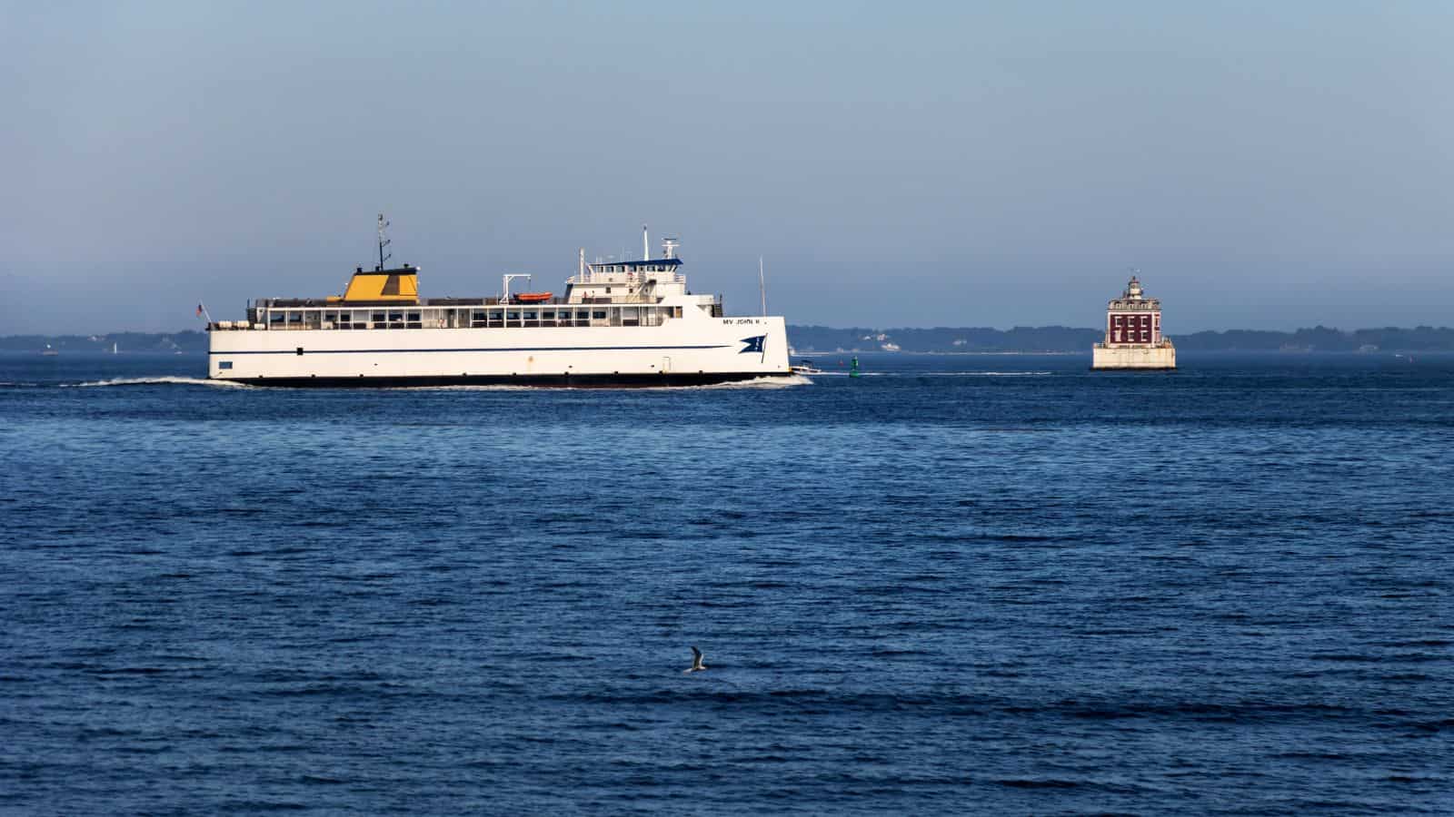 A white ferry travels across a calm body of water near a small lighthouse with land visible in the distant background.