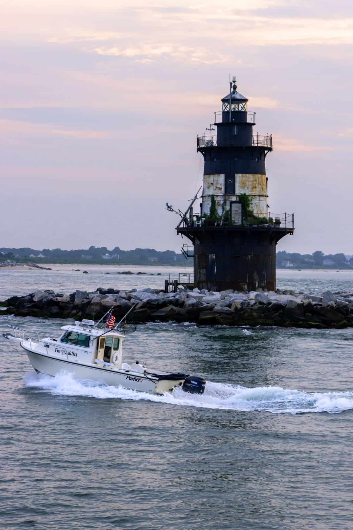 A small motorboat moves past a weathered lighthouse on a rocky breakwater at dusk, with calm water and a distant shoreline in the background.