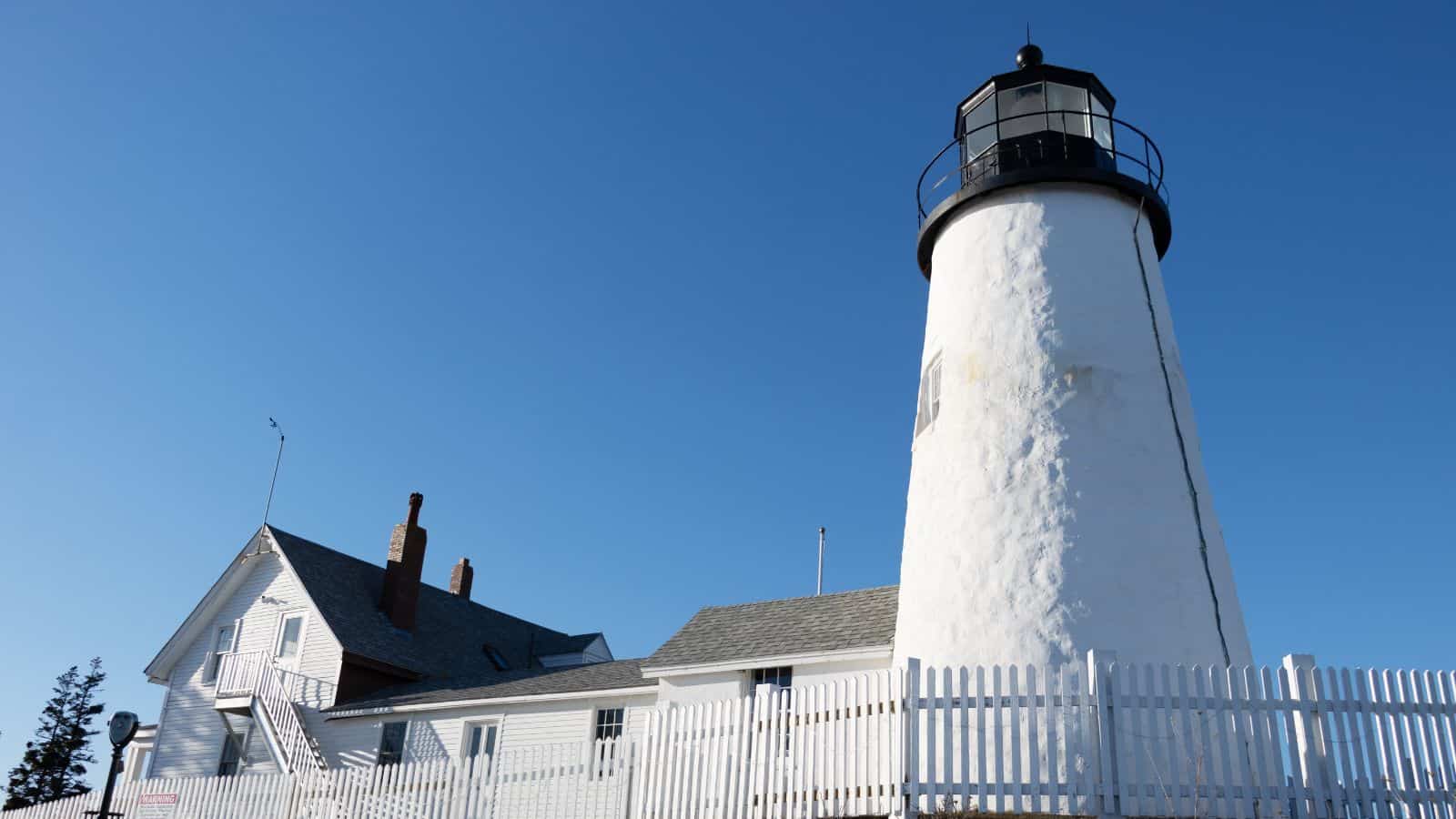 White lighthouse with a black top stands beside a white wooden building, both enclosed by a white picket fence under a clear blue sky.