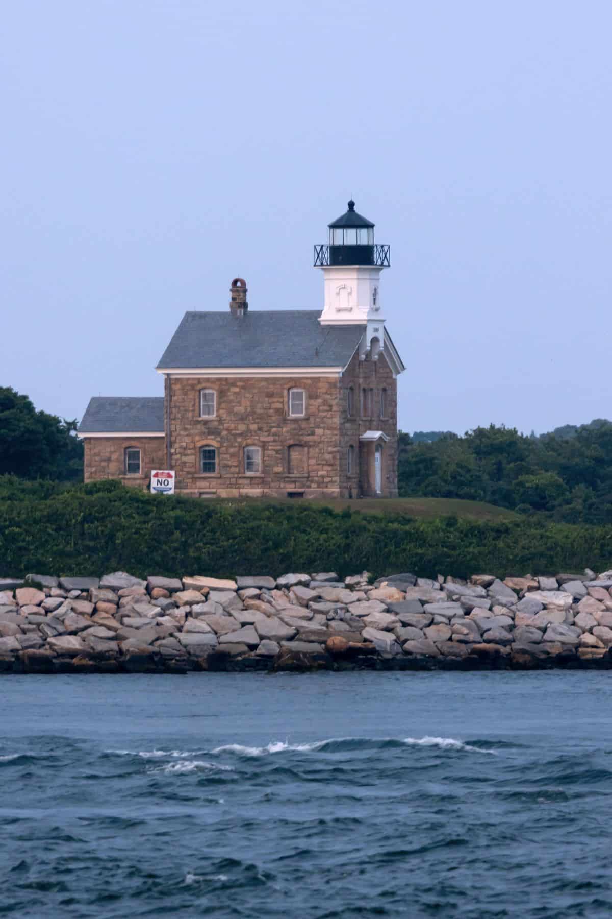 A stone lighthouse with a black lantern stands behind a rocky seawall, surrounded by greenery. Nearby, a "No Trespassing" sign is visible—a scenic view you might spot during a sunset cruise and seal watch with Blackhawk Fishing.