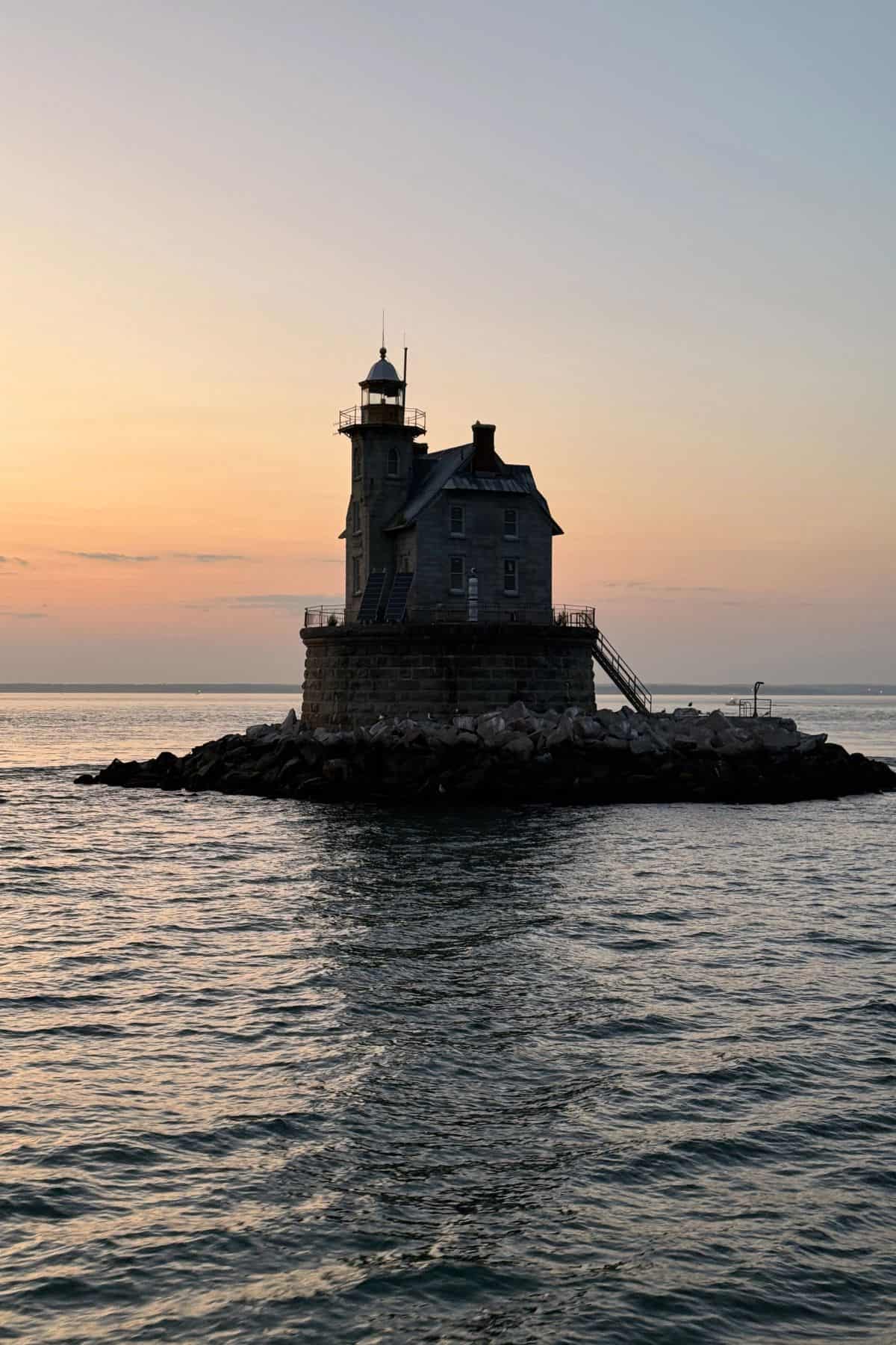 A solitary lighthouse sits on a small rocky island surrounded by calm water at sunset, with a clear sky in the background.