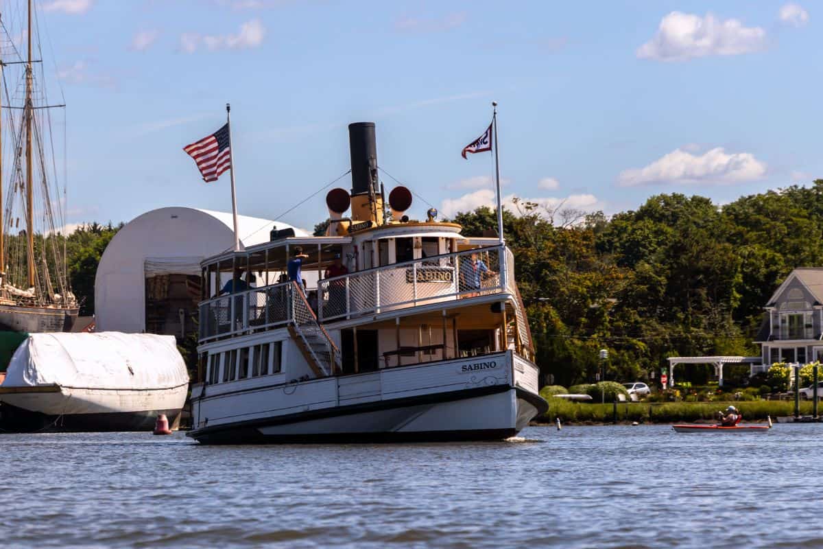 A white paddleboat named Sabino with two flags sails on a river near docks, trees, and a house under a blue sky with scattered clouds.
