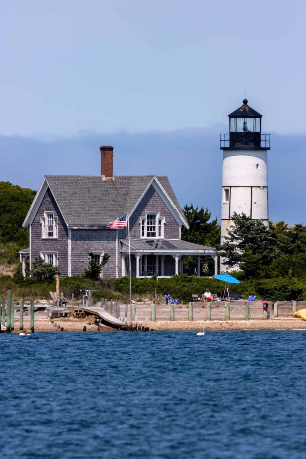 A white lighthouse stands next to a gray shingled house with an American flag, near the shore with people and a wooden dock visible by the water.