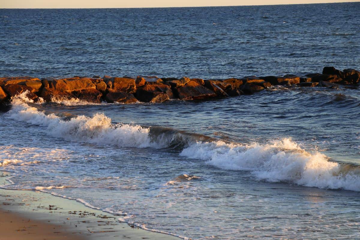 Waves gently breaking on a sandy shore with a stone breakwater in the background and sunlight illuminating the rocks.