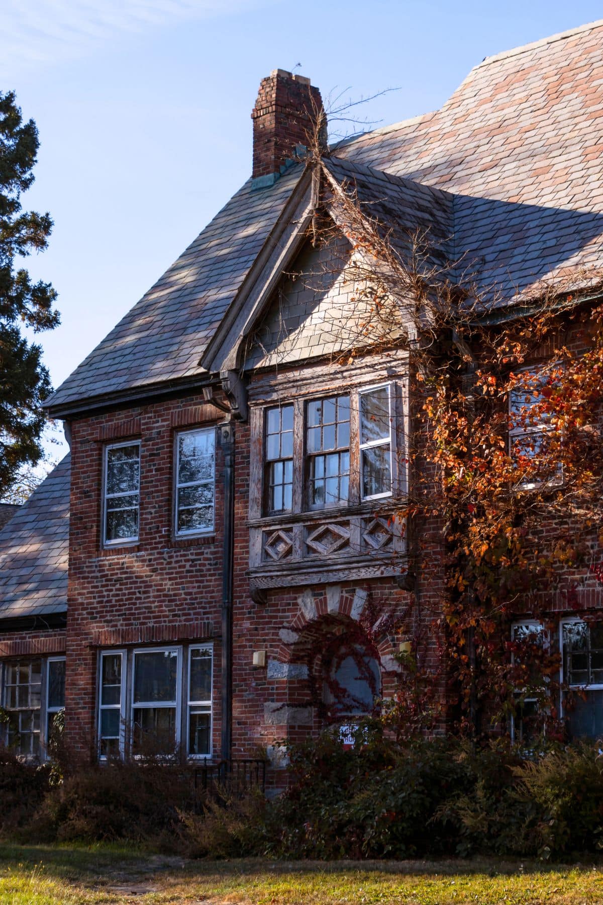 A brick house with a steep slate roof, multiple windows, a prominent chimney, and climbing vines partially covering the exterior.