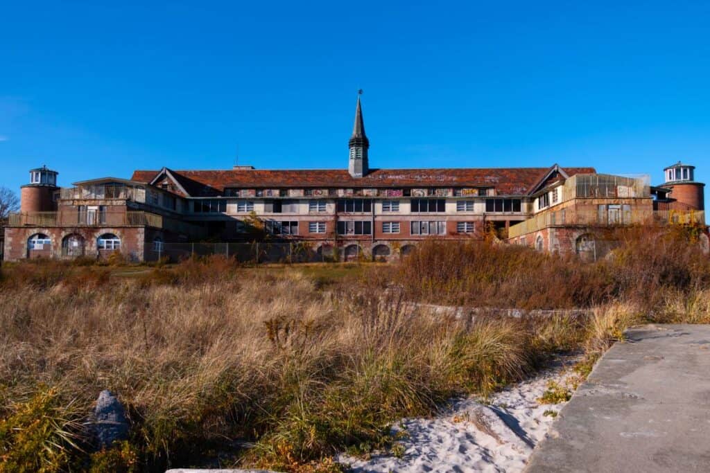 A large, abandoned brick building with a central steeple stands behind overgrown grass and sand under a clear blue sky.