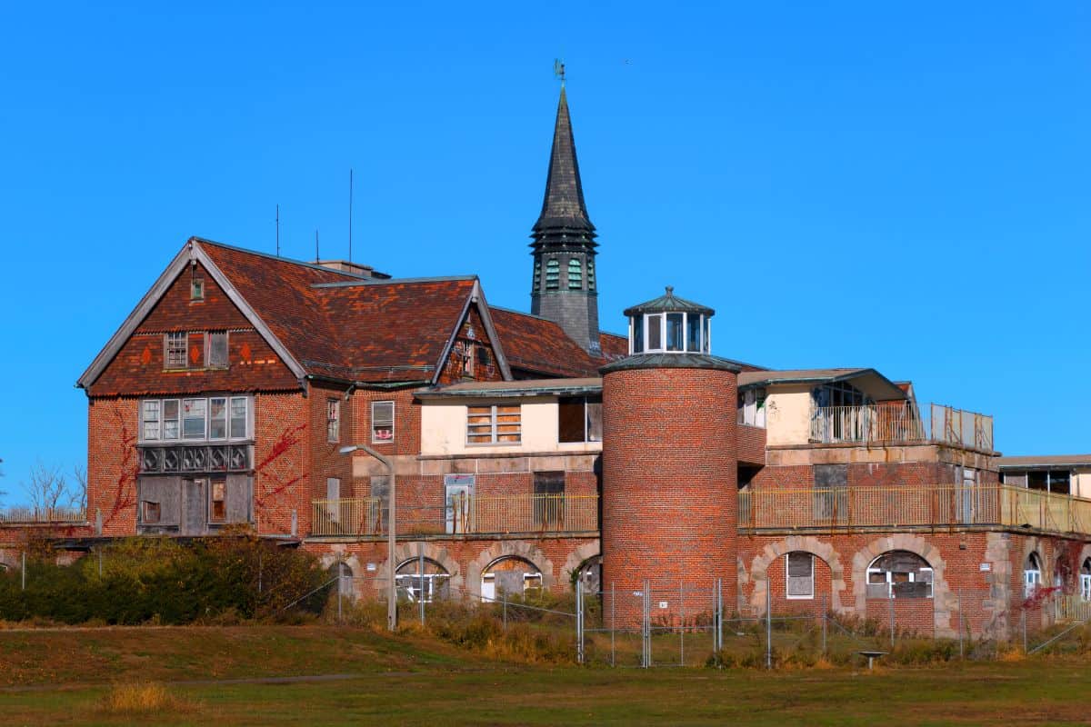 A large, old brick building with boarded windows and a pointed tower stands under a clear blue sky, surrounded by grass and a wire fence.