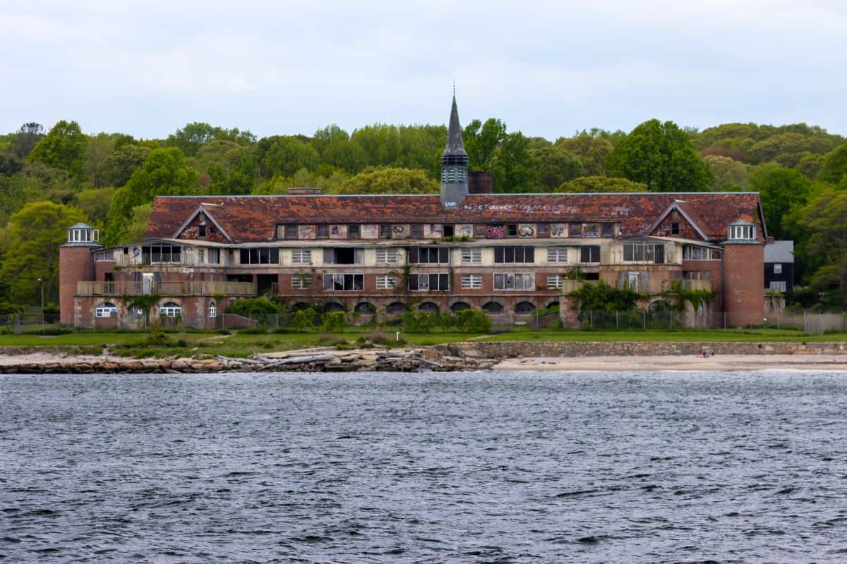 An abandoned, multi-story brick building with boarded windows and graffiti sits near a shoreline, surrounded by trees and vegetation.