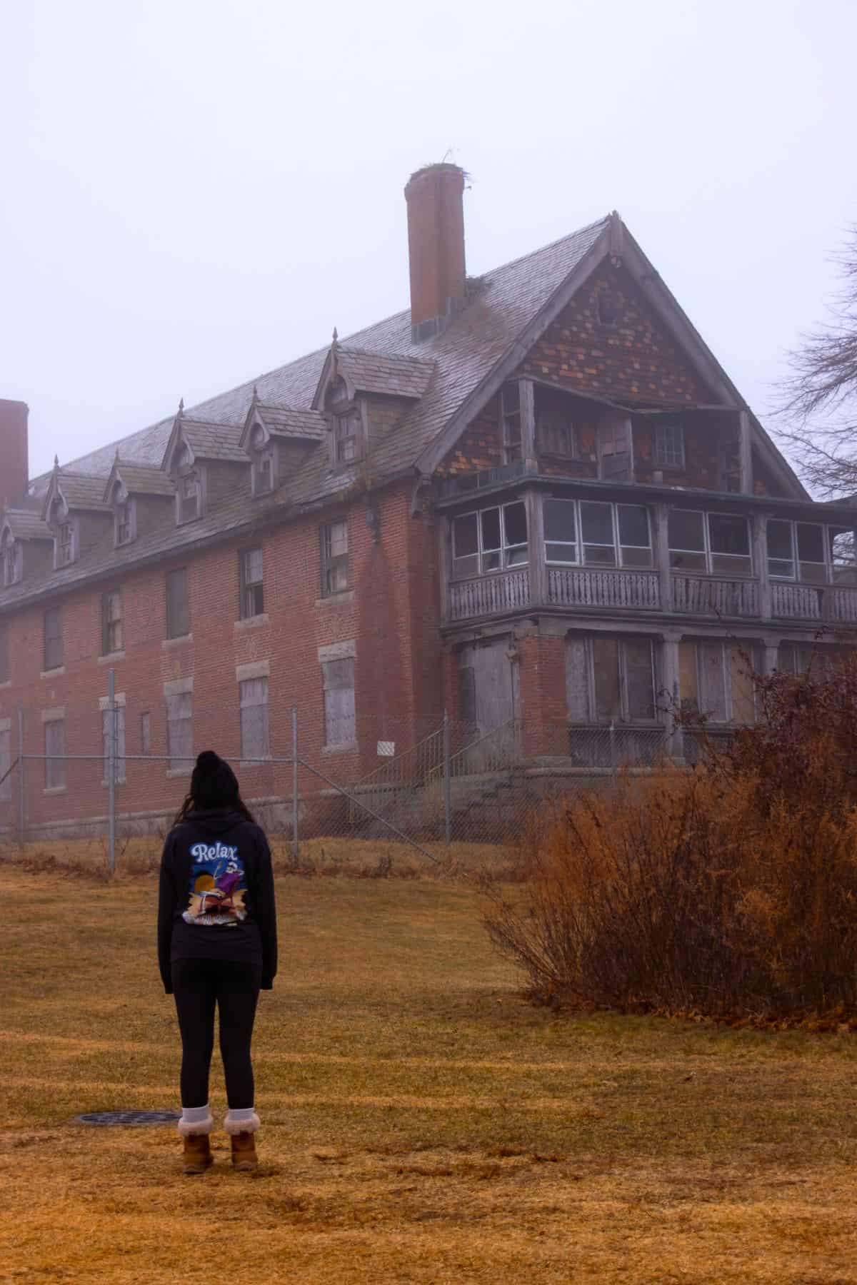 A person stands on a brown grassy lawn facing an old, large, and weathered brick building with boarded windows on a foggy day.