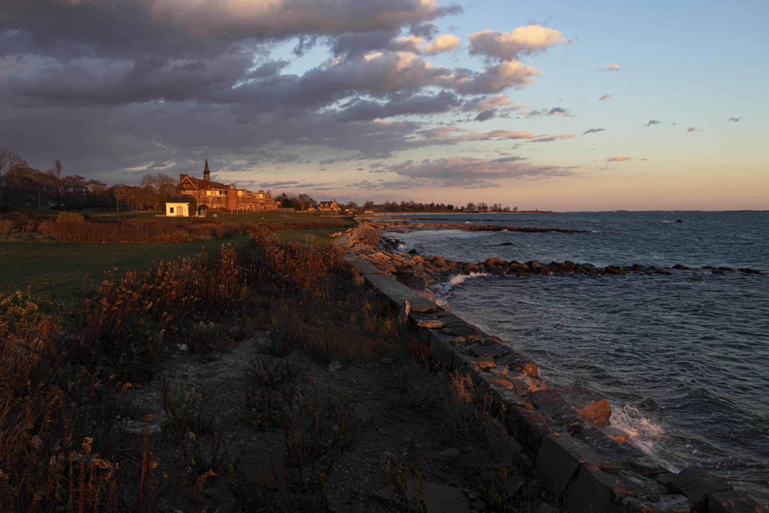 A rocky shoreline at sunset with waves hitting the rocks, grass, and buildings, including a church, visible under a partly cloudy sky.
