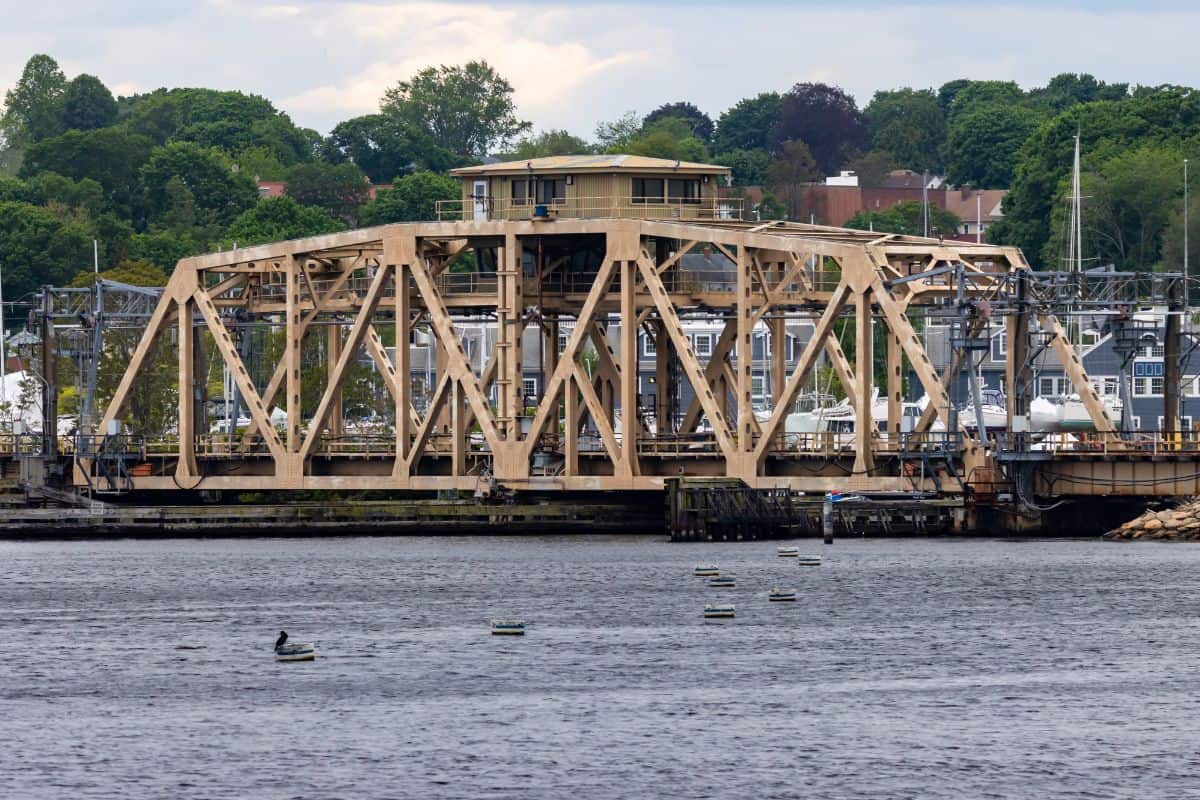 A steel truss drawbridge spans a river, with a control house on top and trees and buildings visible in the background.