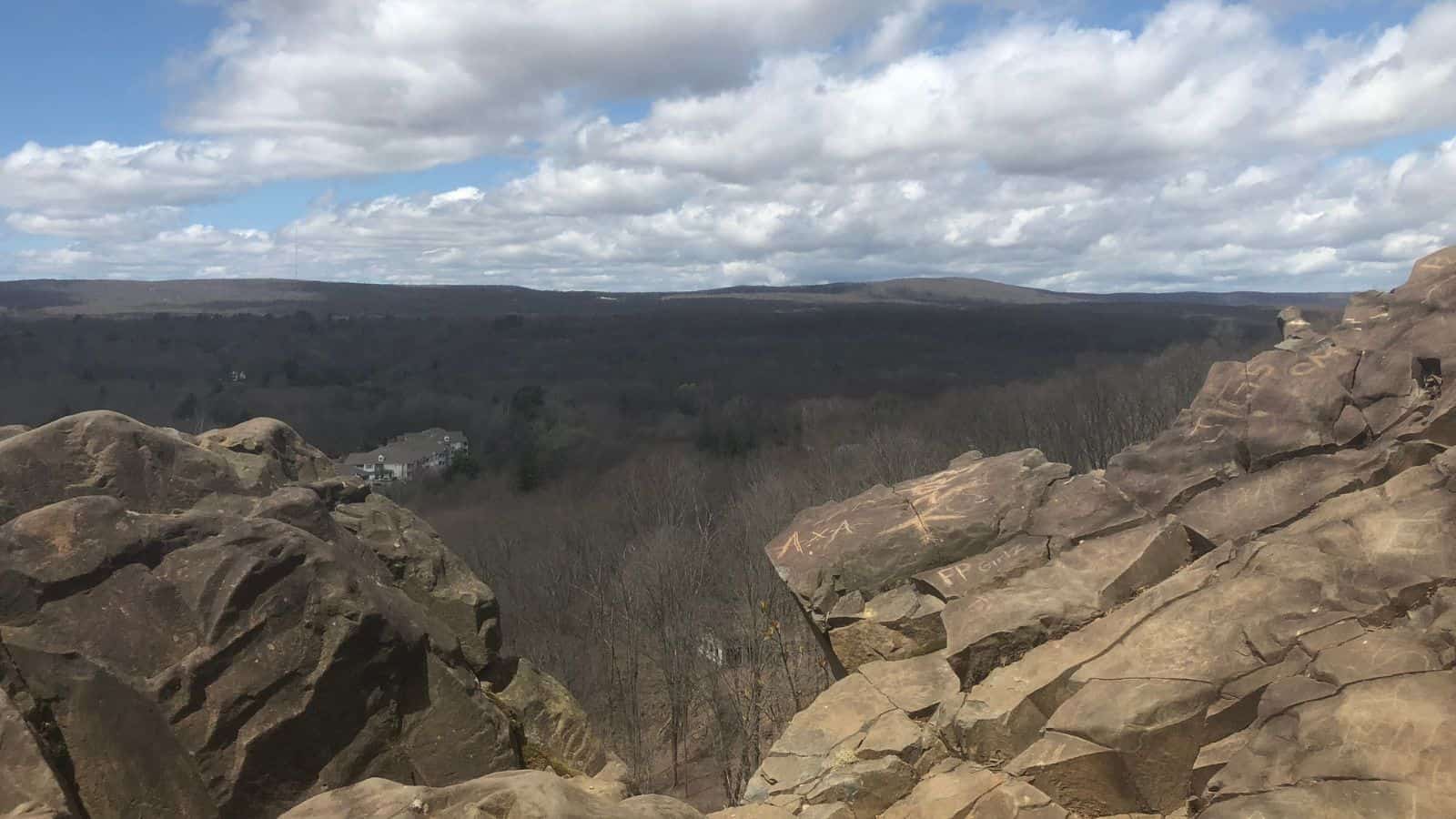A rocky overlook with some graffiti, looking out over a vast forested landscape under a partly cloudy sky.