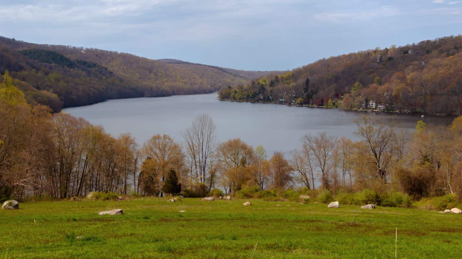 A calm lake bordered by hills with sparse trees, a grassy field with scattered rocks in the foreground, and houses visible along the distant shoreline.