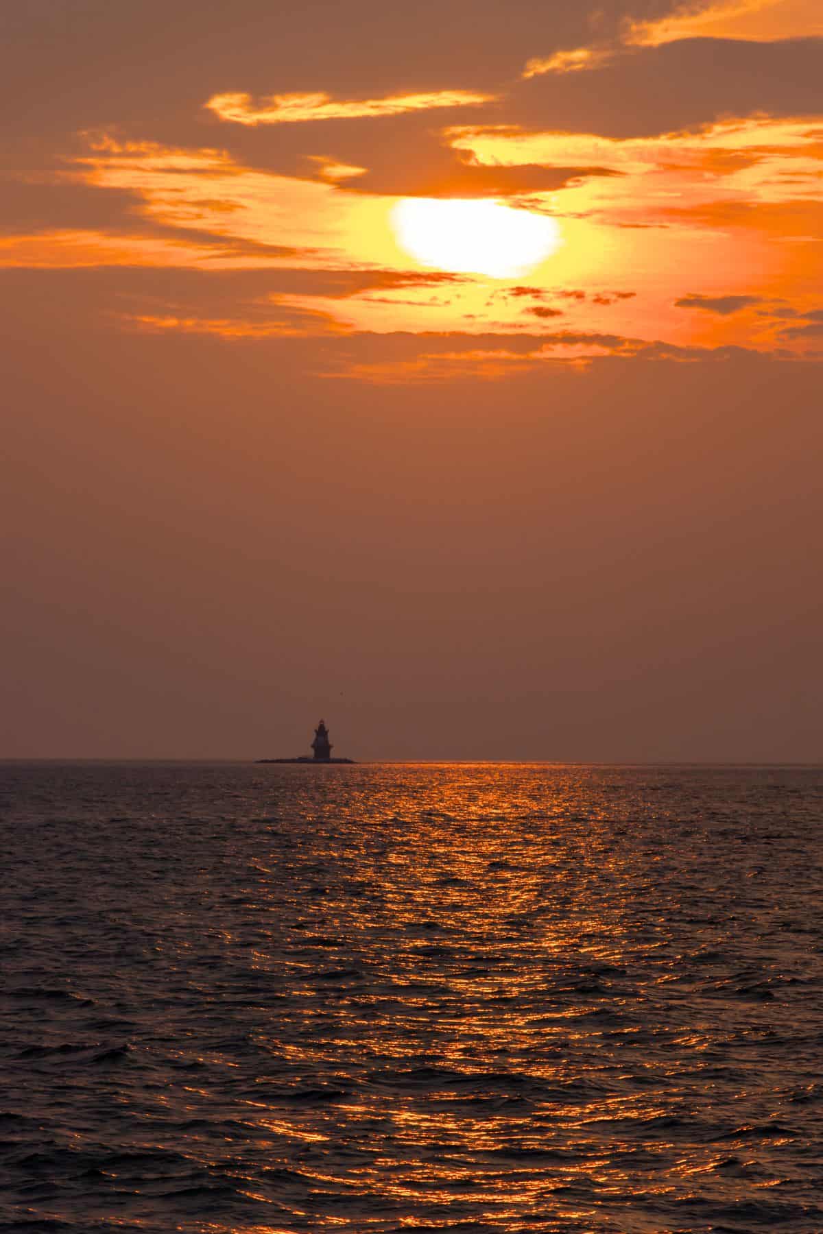 A lighthouse sits on a small island under an orange sunset sky, with golden sunlight reflecting on the calm sea.