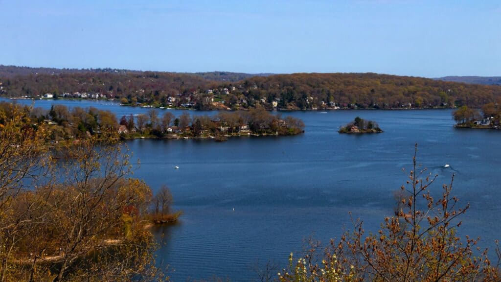 A large lake with several small islands and a shoreline lined with houses, surrounded by trees with early spring foliage under a clear sky.