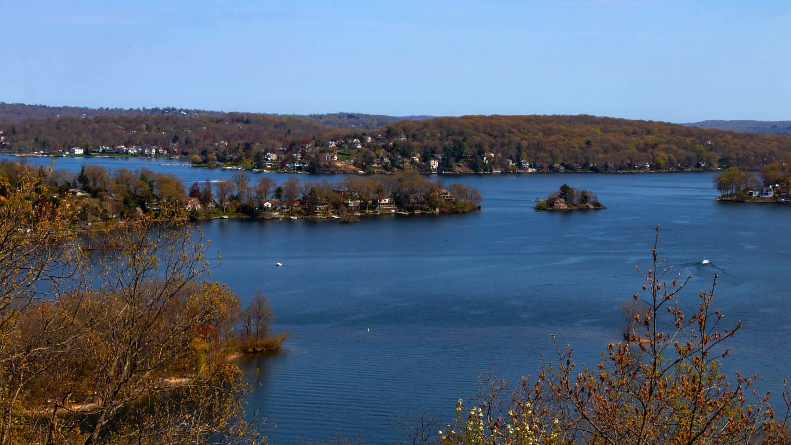 A large lake with several small islands and a shoreline lined with houses, surrounded by trees with early spring foliage under a clear sky.