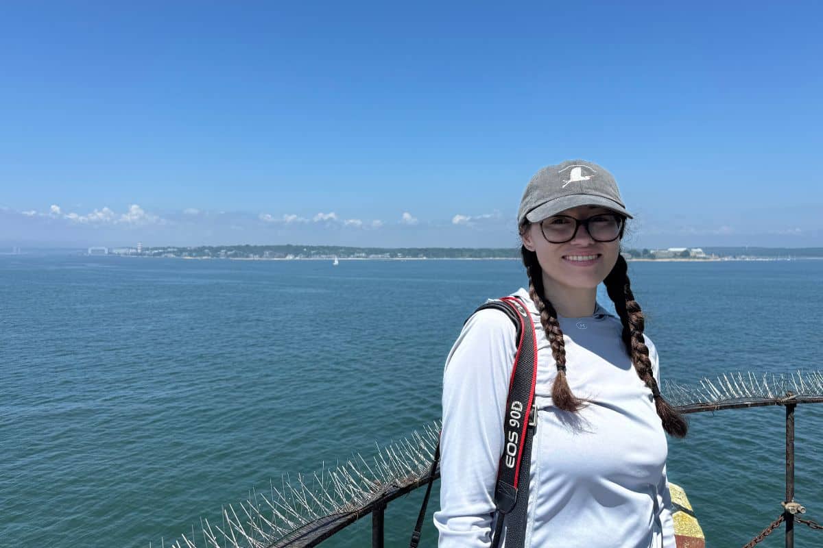 A woman with glasses and braided hair stands on a metal railing overlooking a large body of water, wearing a gray cap and carrying a camera. The distant shoreline is visible under a blue sky.