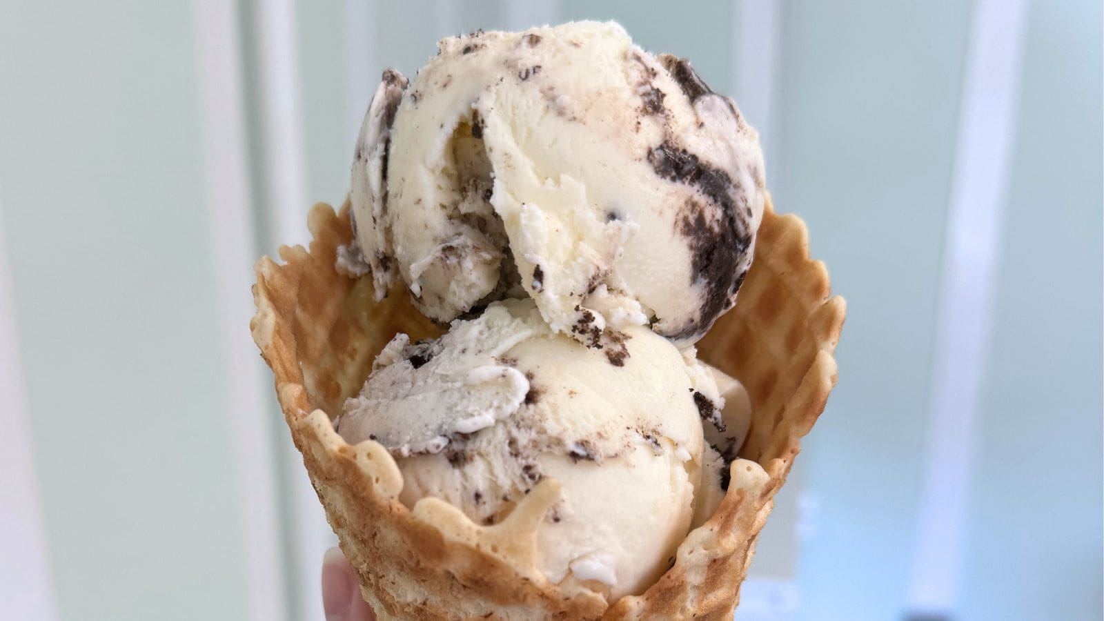 A hand holding a waffle cone with two scoops of cookies and cream ice cream against a light-colored background.