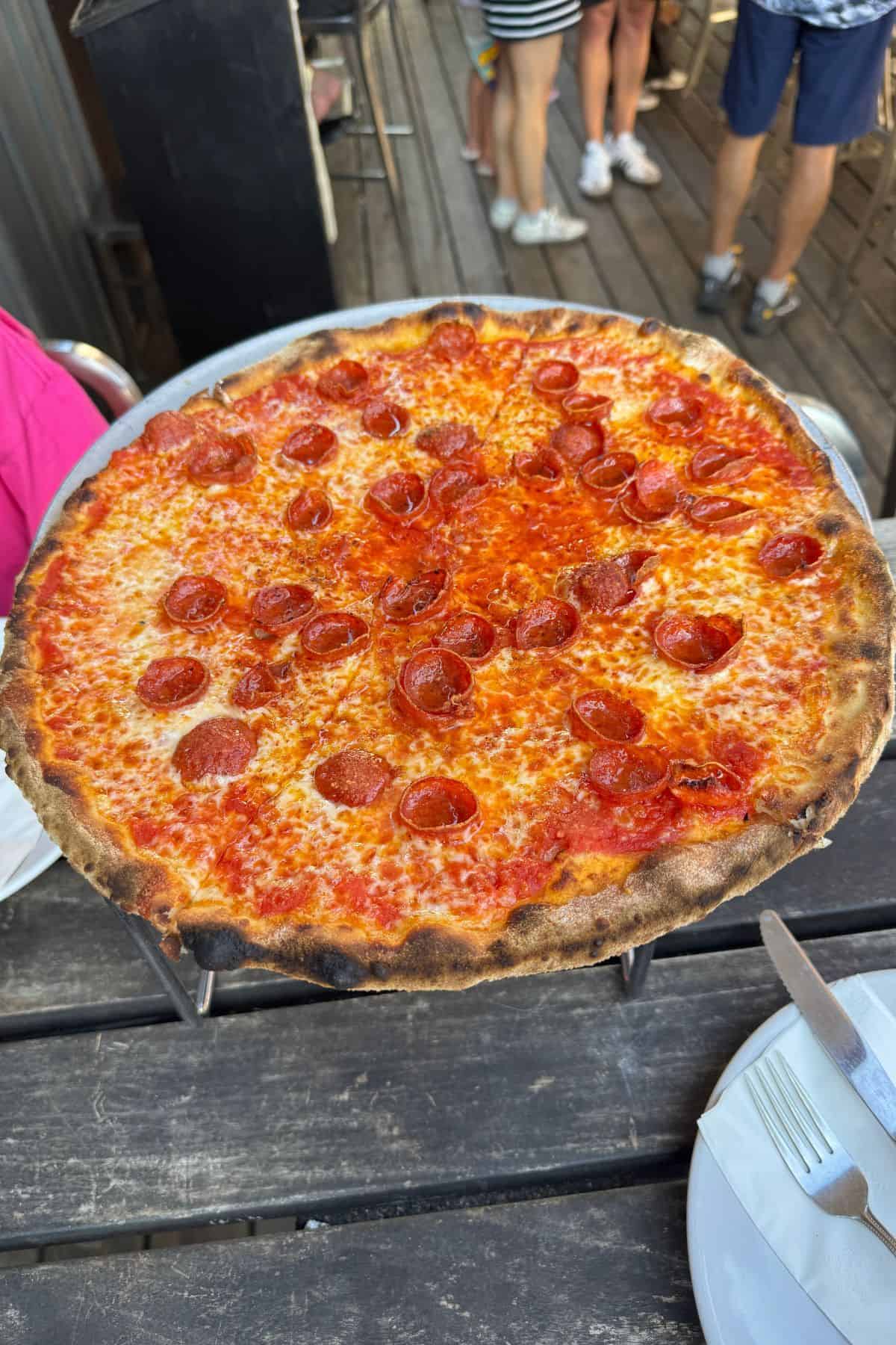 A large pepperoni pizza with a thin, crispy crust is placed on a metal stand above a wooden table set with plates and cutlery. People are visible in the background.