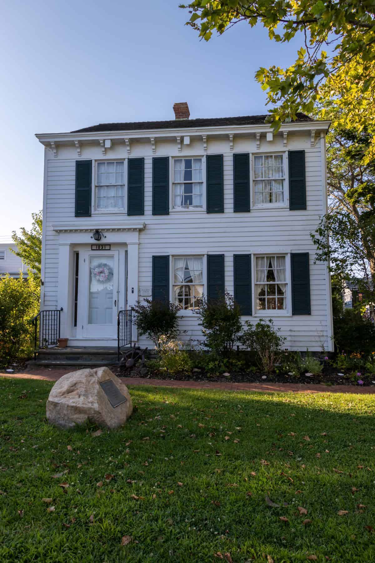 A two-story white house with green shutters, a central front door, and a small garden; a large rock with a plaque sits on the lawn in front.