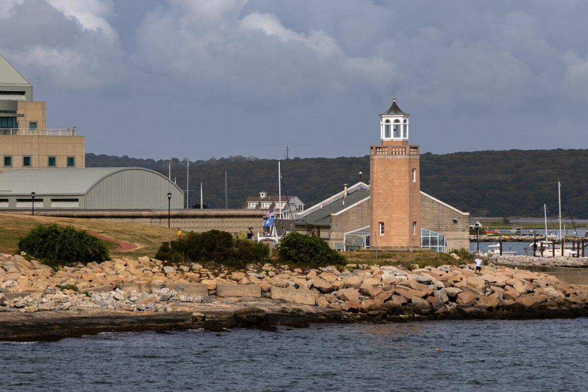 A small lighthouse stands near a stone building on a rocky shoreline with water in the foreground and a forested hill in the background.