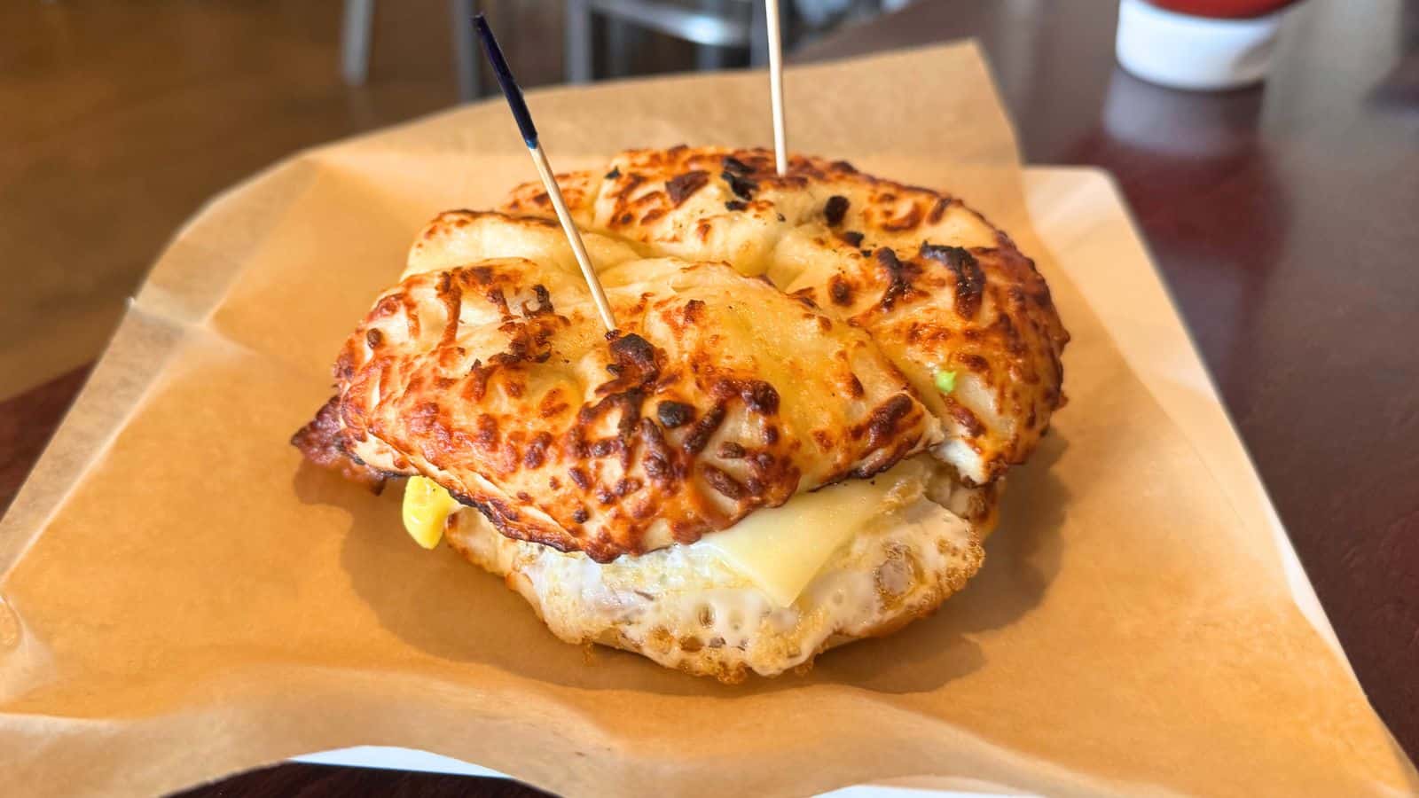 A breakfast sandwich with egg and cheese on a cheese bagel is secured with two toothpicks and served on brown parchment paper.