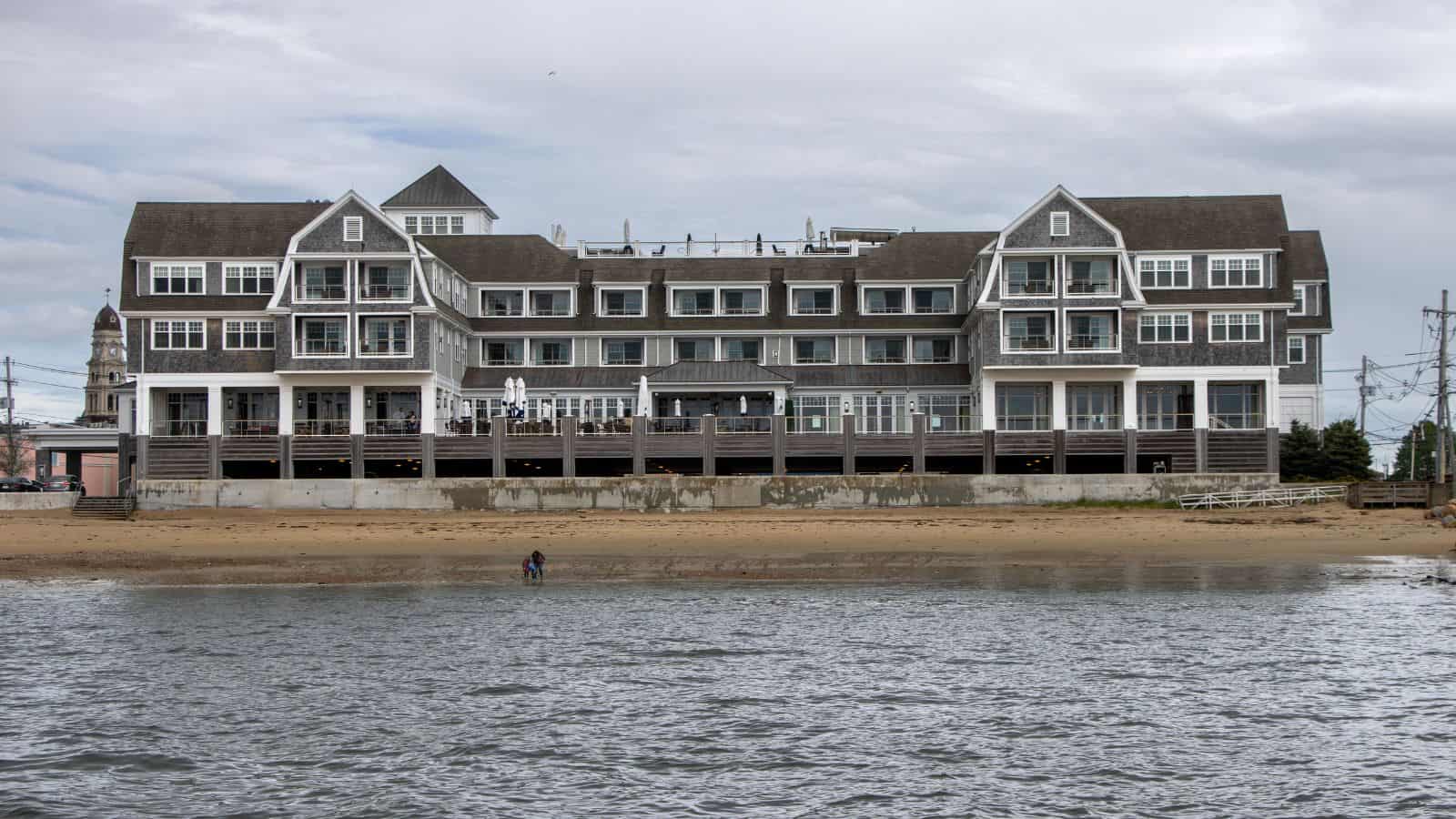 A large, multi-story waterfront building with balconies overlooks a sandy beach, with two people standing near the water under a cloudy sky.