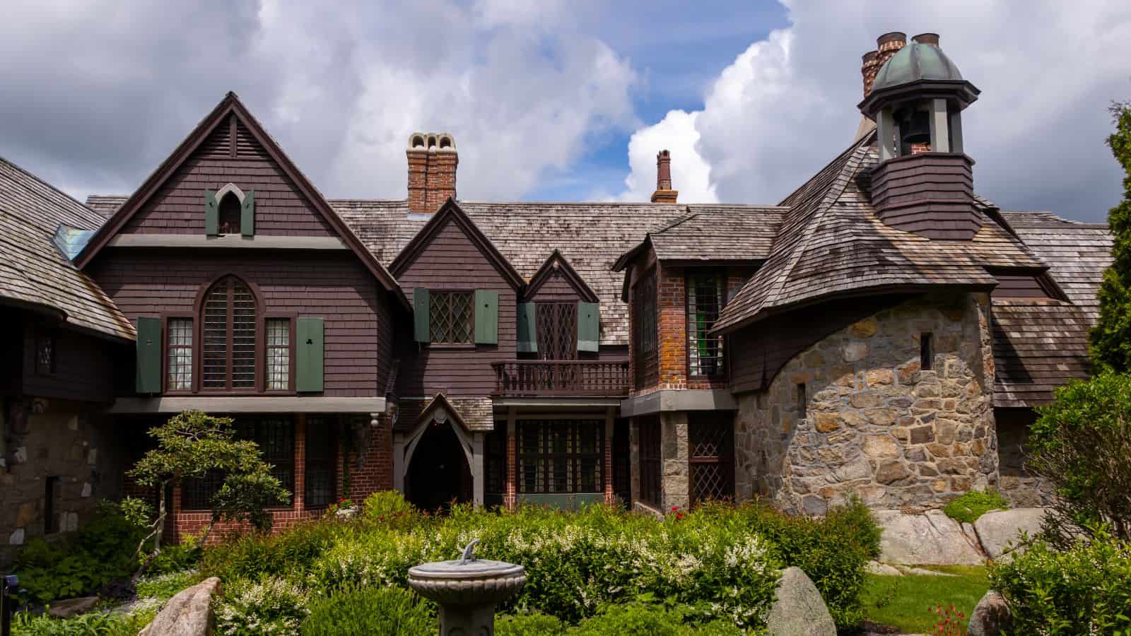 Large, historic stone and wood house with steep gabled roofs, arched windows, and a landscaped garden in front under a cloudy sky.