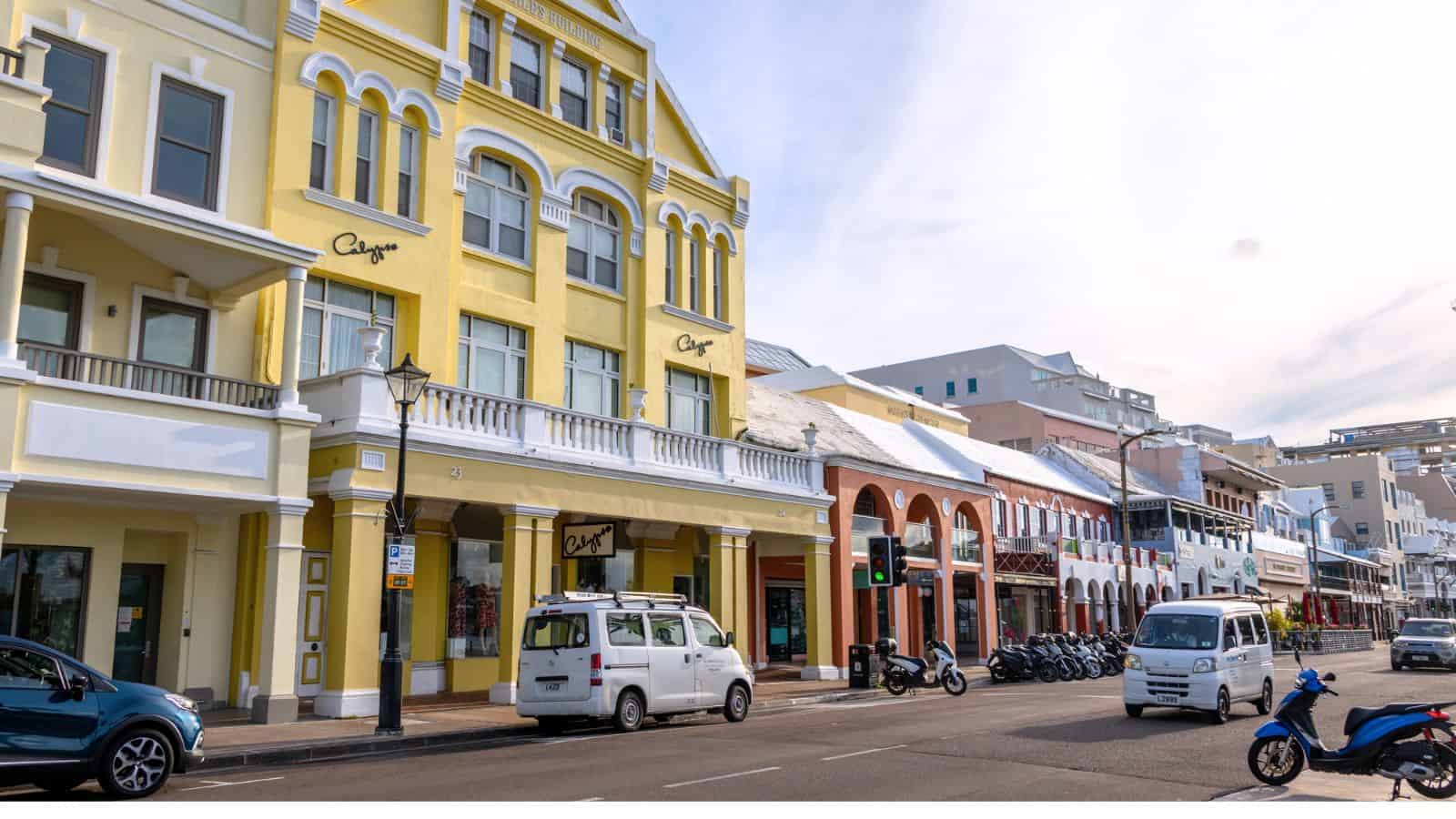 A street scene with yellow and red buildings, parked cars, motorcycles, and a clear sky in an urban area.