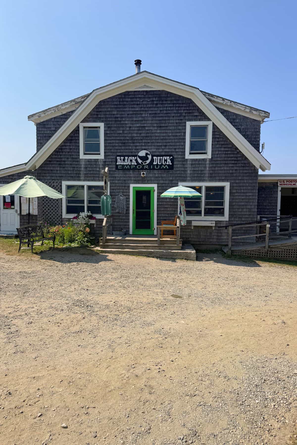 A rustic wooden building on Monhegan Island with a sign reading "Black Duck Emporium," featuring green double doors, outdoor seating, and umbrellas on a sunny day.