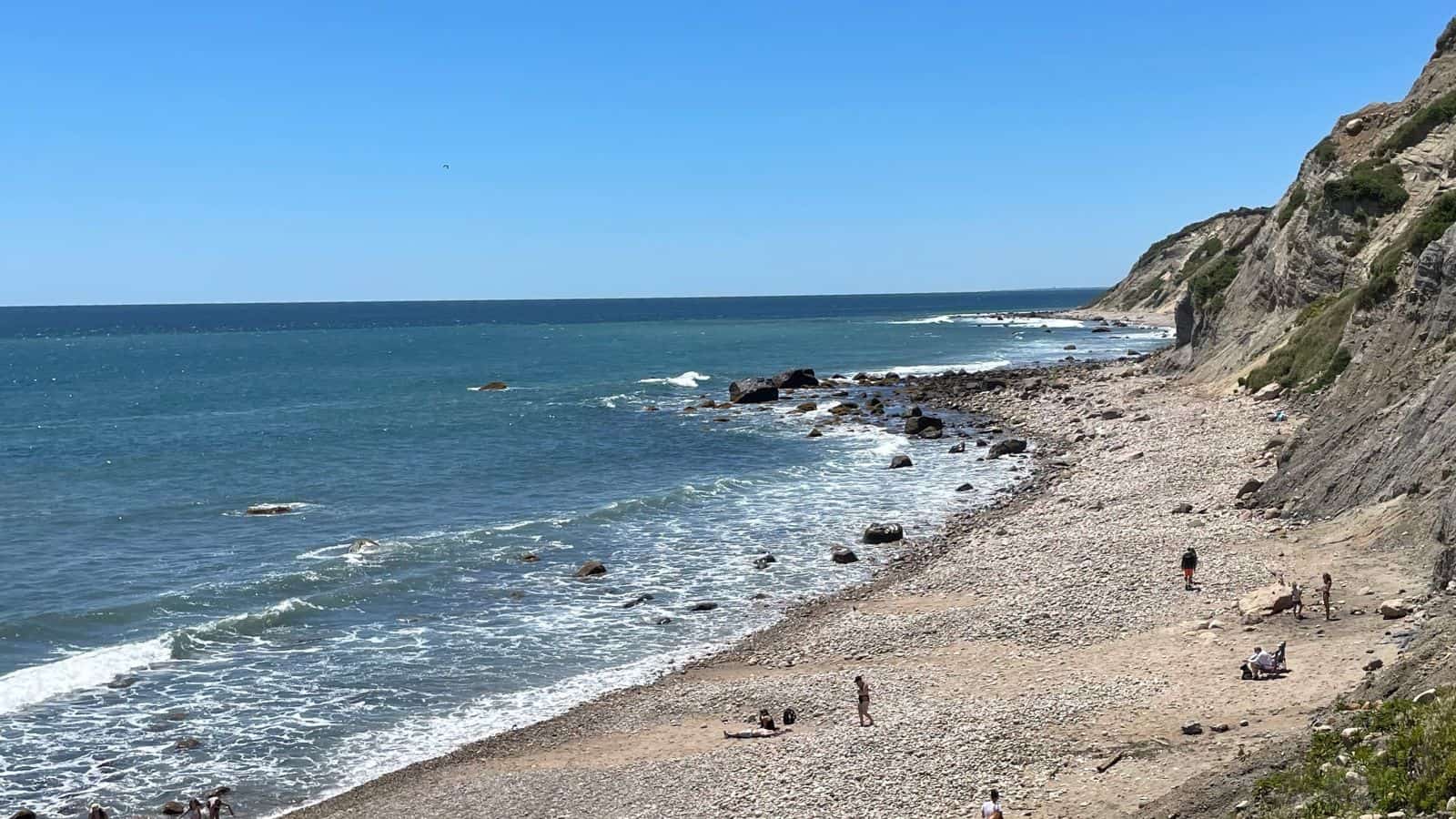 Rocky shoreline with scattered people along the beach, waves approaching, and cliffs rising to the right under a clear blue sky.
