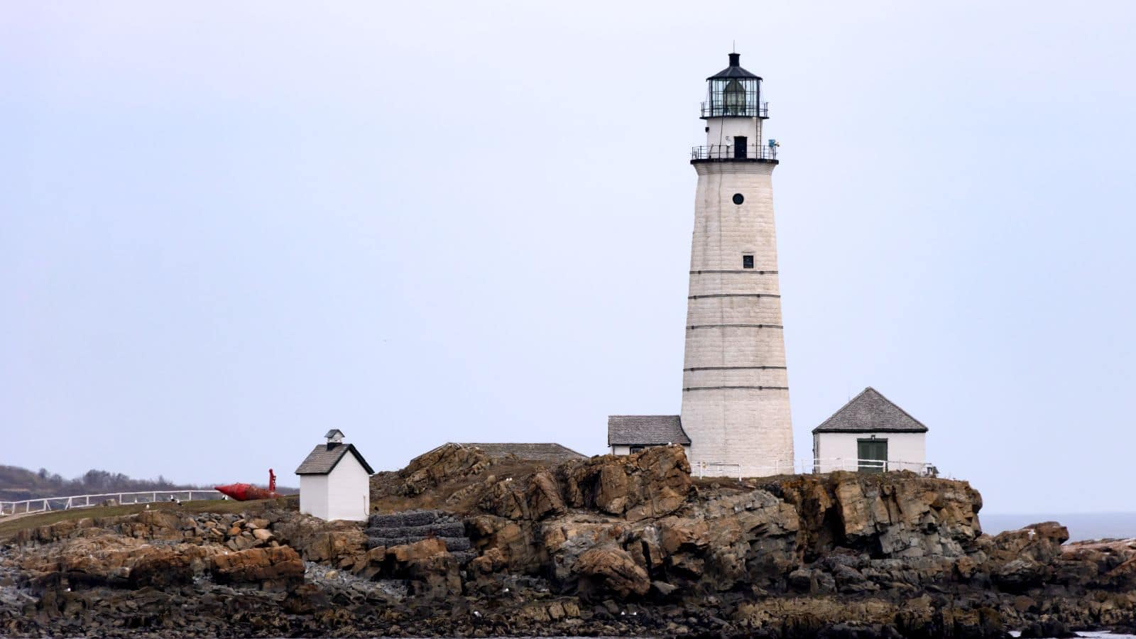 A white lighthouse with adjacent small buildings stands on rocky terrain by the water under an overcast sky.