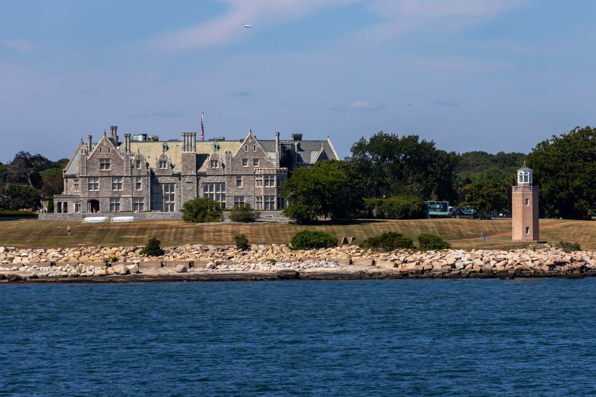 Large stone mansion with multiple gables sits on a grassy lawn near the shoreline; a small lighthouse stands on the right beside a rocky barrier.
