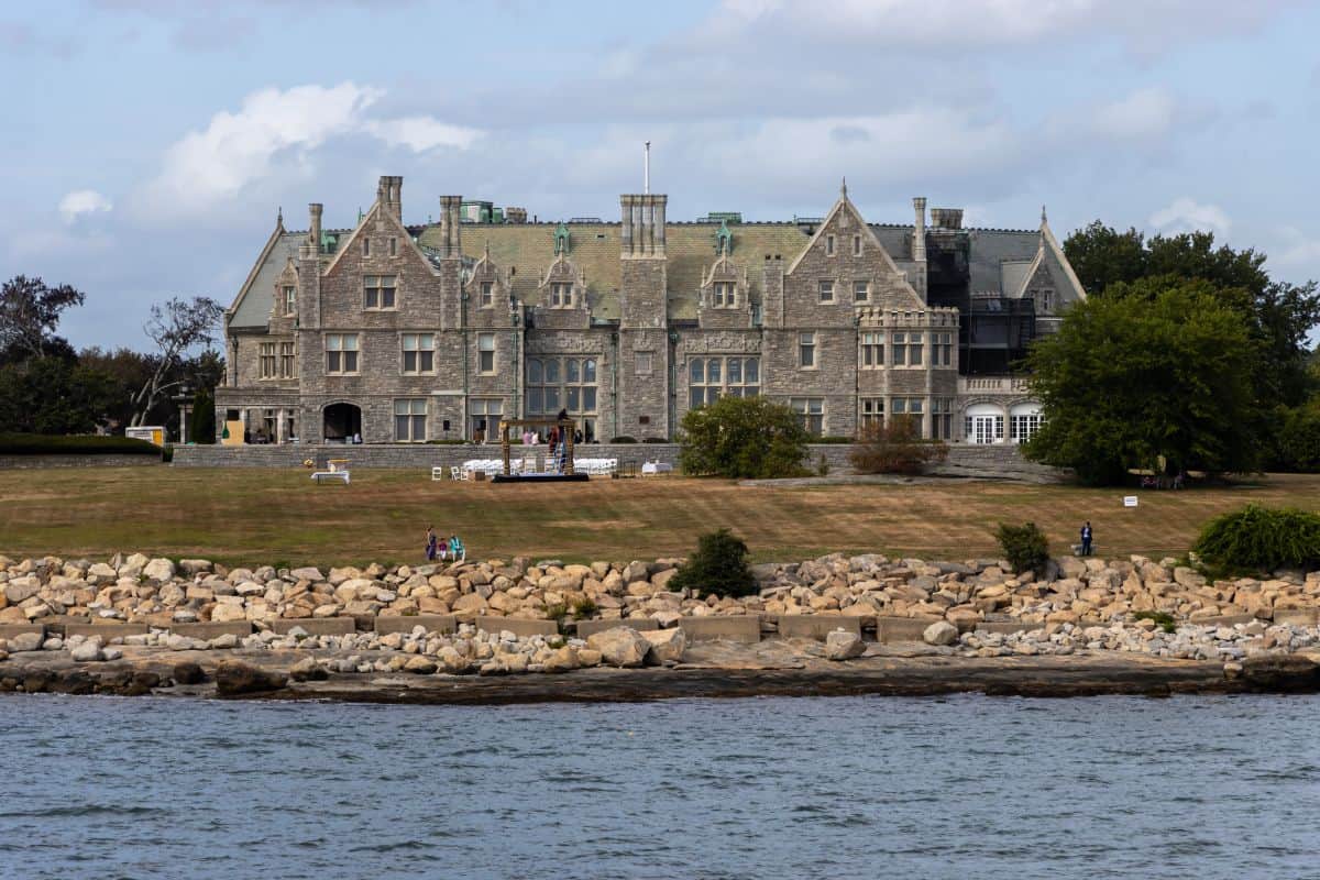 A large stone mansion with steep roofs and many chimneys stands on a grassy hill by the water, with rocks lining the shore and trees in the background.