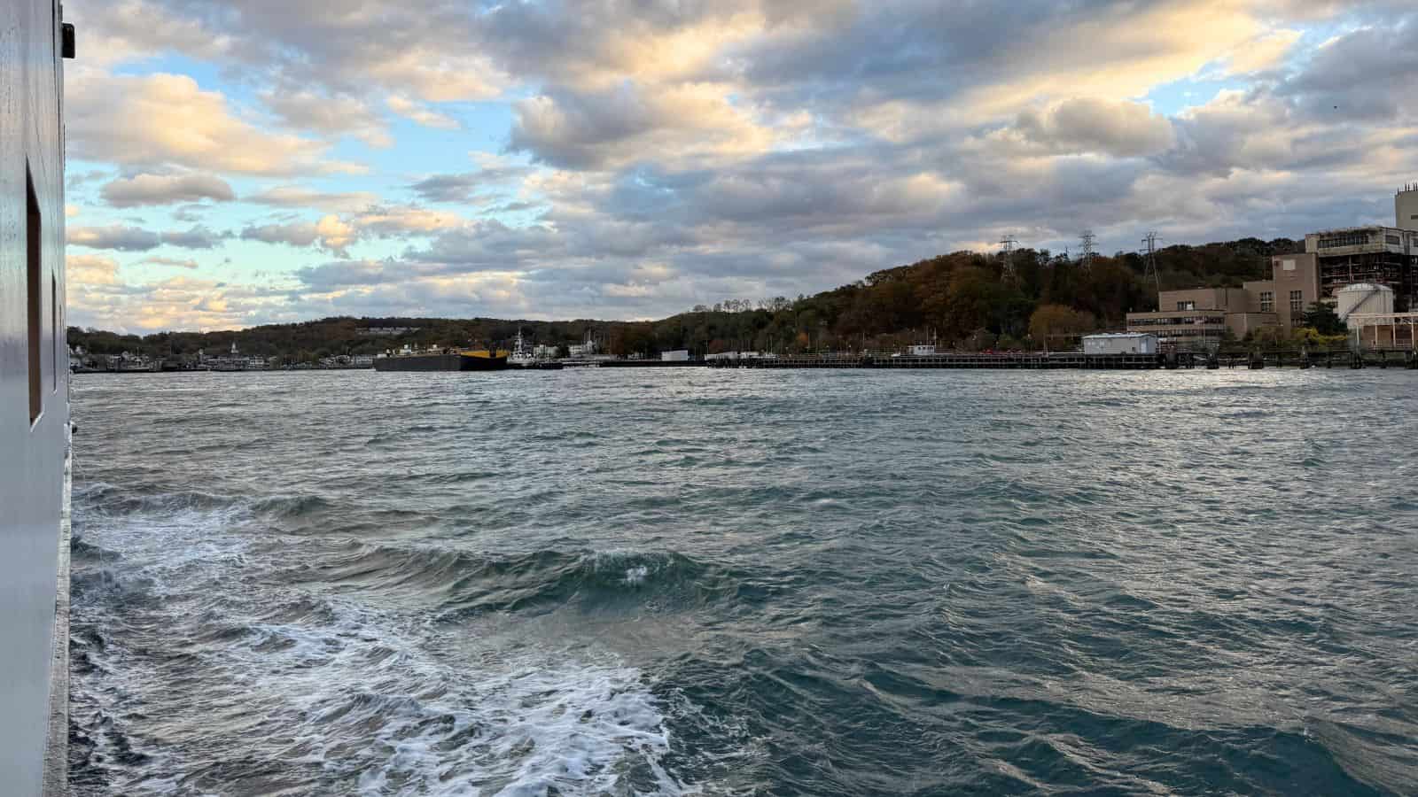 A view from a boat ride to in New England reveals choppy water, a shoreline dotted with buildings and trees, and a partly cloudy sky at sunset.