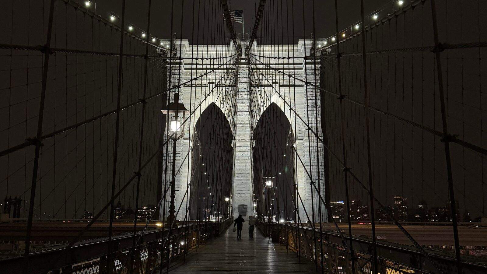 The Brooklyn Bridge at night, illuminated with lights, with a few people walking along the pedestrian pathway and city buildings visible in the background.