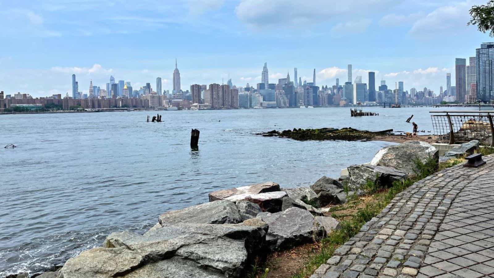 View of Manhattan skyline across the East River, seen from a rocky shoreline and cobblestone path on the Brooklyn side under a partly cloudy sky.