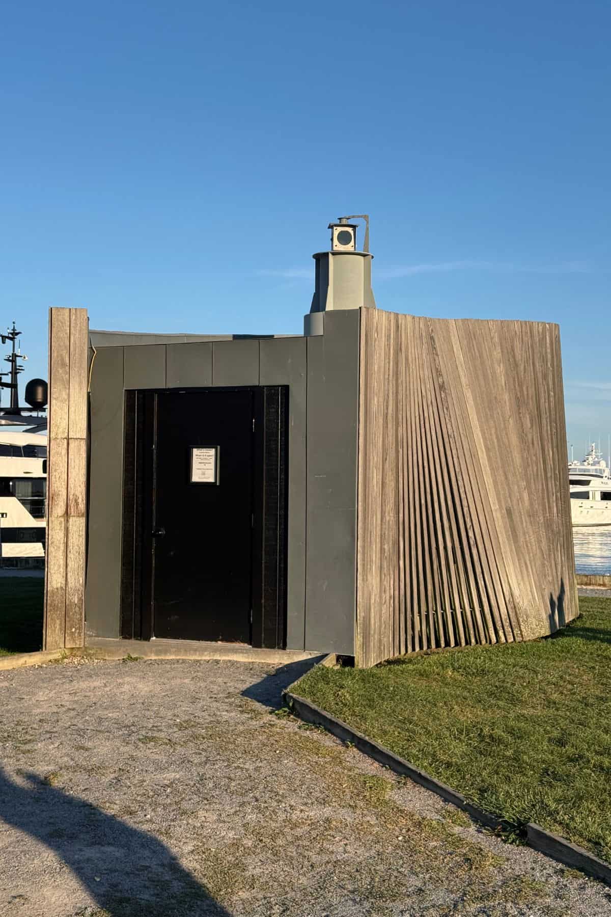 A small, modern building with a black door, a slatted wooden panel, and a notice on the door, situated on a grassy area near a marina.