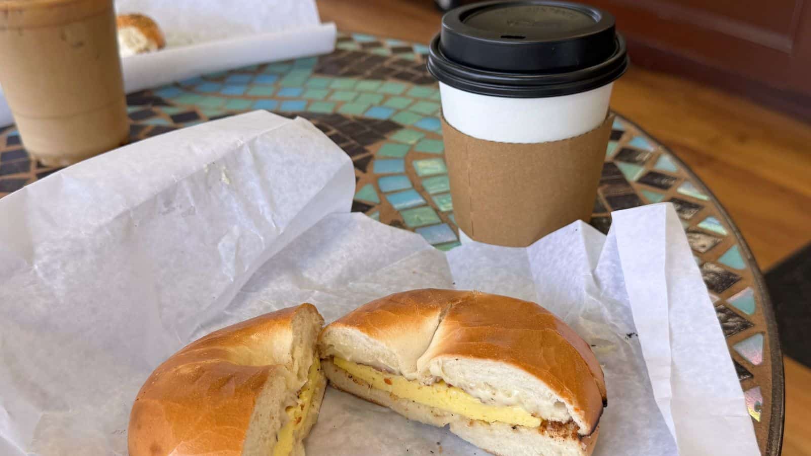 A breakfast sandwich with egg and cheese on a bagel sits on parchment paper next to a hot coffee in a to-go cup on a mosaic table.