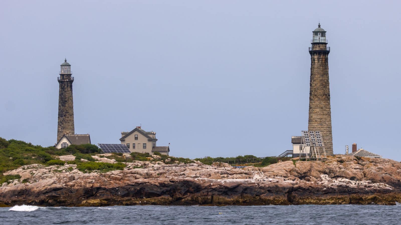 Two tall stone lighthouses stand on a rocky coastline with several small buildings and greenery under a cloudy sky.