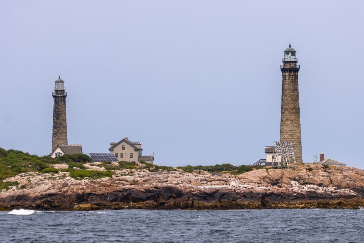 Two tall stone lighthouses stand on a rocky shoreline with a few houses and greenery under a cloudy sky, viewed from the water.