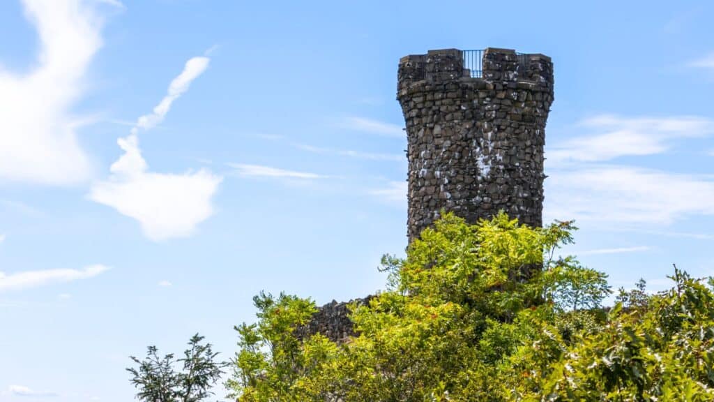 Stone observation tower with a crenellated top rises above green foliage against a partly cloudy blue sky.