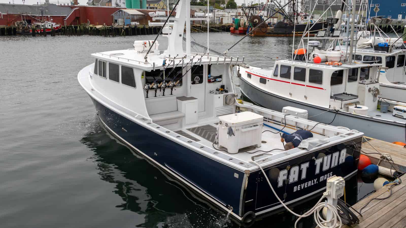 A white and blue fishing boat named "FAT TUNA" docked at a marina, with fishing gear visible and a person working near the back of the boat.