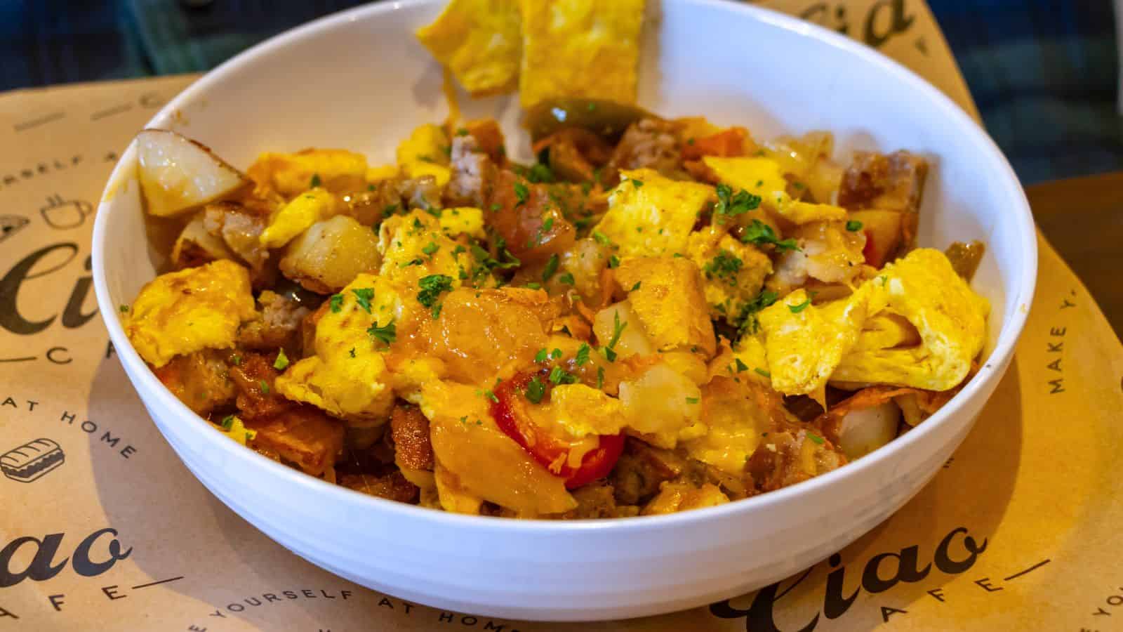 A white bowl filled with scrambled eggs, potatoes, onions, and bell peppers, garnished with chopped herbs, evokes the hearty fare found at the best breakfast places in Connecticut, and sits on a brown paper placemat.