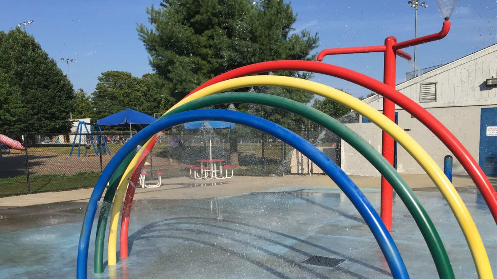 A splash pad with red, yellow, green, and blue curved water spray arches, picnic tables, and a playground in the background.